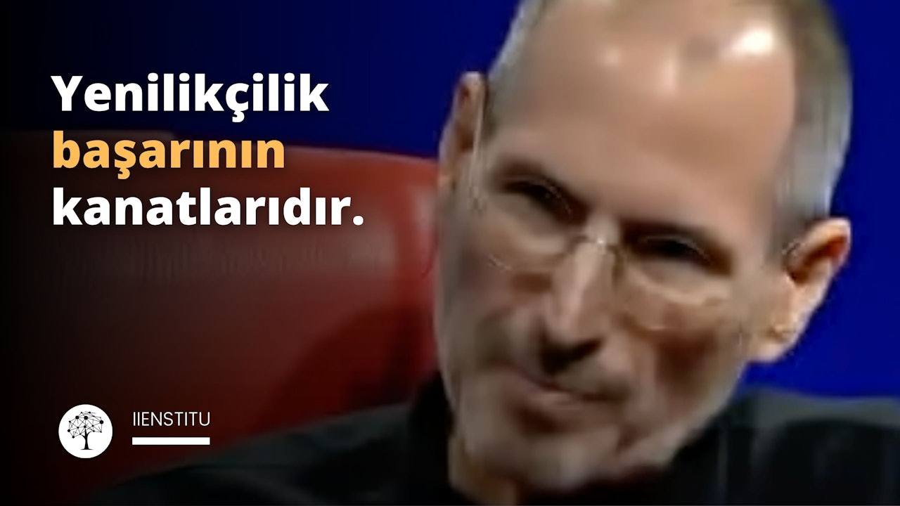 Kare, siyah çerçeveli camları olan bir gözlük ve siyah bir tişört giyen bir adam kırmızı bir sandalyenin önünde durmaktadır. Kollarını kavuşturmuş, bıyıklı ve yüzünde ciddi bir ifade var. Arka planda, üzerinde beyaz yazılar olan mavi bir duvar, siyah ve beyaz şekillerden oluşan bir logo ve siyah zemin üzerinde sarı bir harf var. Ayrıca, uzakta bir kişinin yüzünün bulanık bir yakın çekimi var. Adamın gözlükleri ve tişörtü arka planda göze çarpıyor ve onu görüntünün odağı haline getiriyor.