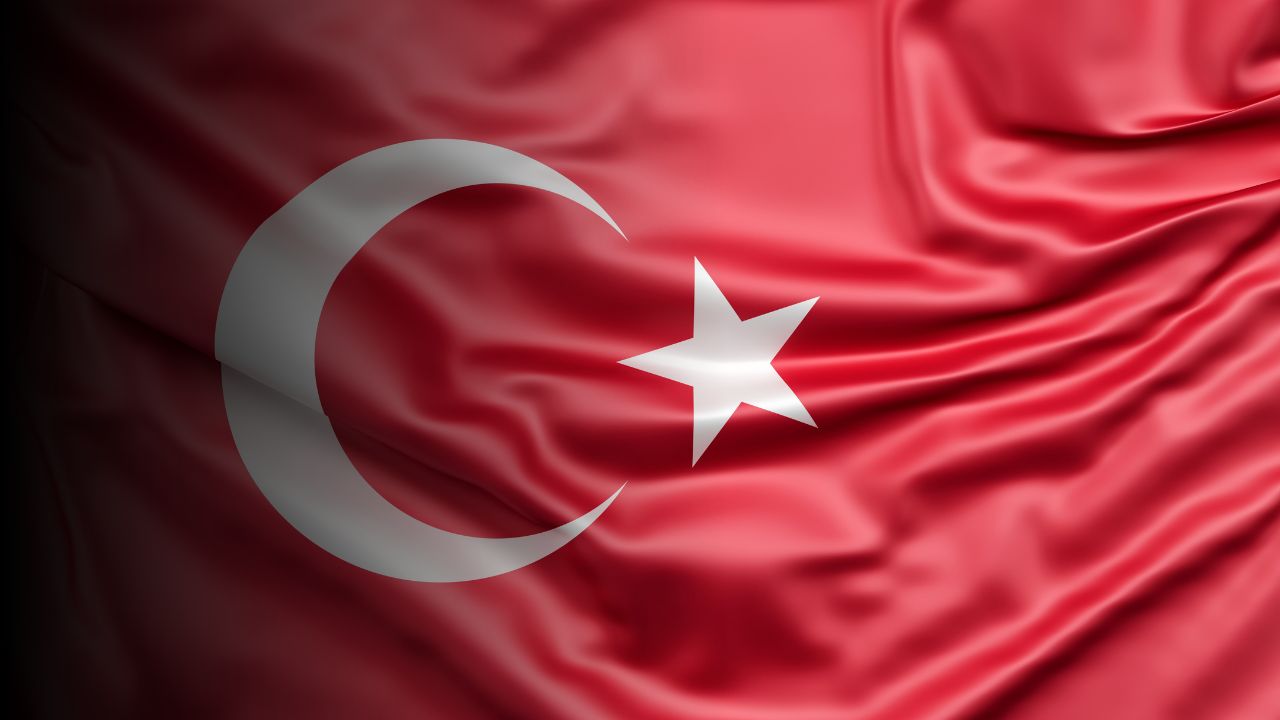 Bu görsel, ortasında beyaz bir hilal ve bir yıldız bulunan canlı kırmızı bir bayrağı göstermektedir. Hilal yarım daire şeklinde kavislidir ve yıldız hilalin ortasındadır. Bayrak resmin sol üst köşesine yerleştirilmiş ve büyük bir kısmı görülebiliyor. Bayrak dikdörtgen bir şekle sahiptir ve parlak kırmızı bir renk tonundan oluşmaktadır. Beyaz hilal ve yıldız resmin ana unsurlarıdır ve kırmızı arka plana karşı öne çıkarlar. Yıldızın sekiz noktası vardır ve hilal ince beyaz çizgilerle belirtilmiştir. Bayrak beyaz bir arka plan üzerinde duruyor ve görselde başka hiçbir nesne ya da özellik bulunmuyor.