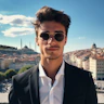 Bu görüntüde güneş gözlüklü ve takım elbiseli bir adam açık havada bir binanın önünde dururken görülüyor. Gülümsüyor ve elleri cebinde, yana doğru bakıyor. Saçları koyu kahverengi ve düzgünce şekillendirilmiş ve çenesinde sakal var. Beyaz bir gömlek üzerine siyah bir takım elbise ceketi, siyah pantolon ve ayakkabı giyiyor. Bu kıyafetin üzerine yansıtıcı gümüş renkli havacı tarzı bir güneş gözlüğü takıyor. Arkasındaki gökyüzü mavi ve bulutlar dağılmış durumda, altındaki zemin ise çimenlik alanlarla çevrili beton kaldırımlardan oluşuyor. Arka planda dağların yanı sıra yakınlardaki diğer binalar da görülebiliyor.