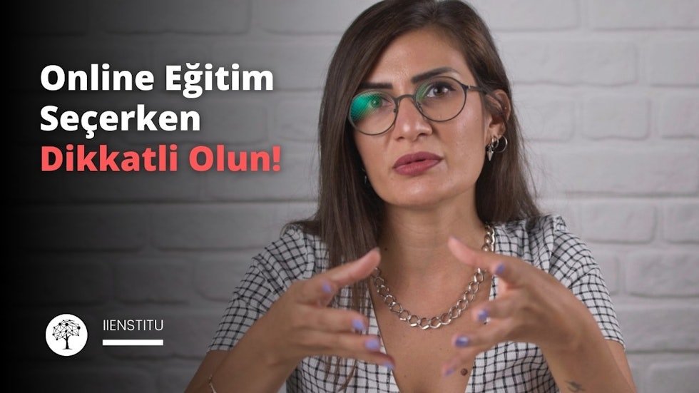 Görselde, beyaz tuğla duvar önünde duran genç bir kadın gözükmektedir. Kadın, kahverengi saçlı, gözlük takmış, ve şık bir kıyafetle (kontrol desenli ceket) giyinmiştir. Kadının yüz ifadesi ciddi ve konuşurken el jestleri kullanmaktadır. Görselin üst kısmında büyük kırmızı harflerle "Online Eğitim Seçerken Dikkatli Olun!" yazmaktadır. Görselin alt sol köşesinde ise beyaz bir ağaç logosu bulunmaktadır ve logo altında "İİENSTITU" yazmaktadır. Bu görsel, bir online eğitim konulu video veya sunumun başlangıcını ya da tanıtımını temsil ediyor gibi görünmektedir.