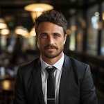 Giovanni Ricci olduğu tahmin edilen, kravatlı klasik siyah takım elbise giymiş bir adamın görüntüsü. Koyu renk, düzgünce şekillendirilmiş saçları ve bakımlı bir sakalı var ve kendinden emin bir gülümsemesi var. Ortam, muhtemelen bir kafe veya restoran gibi sofistike bir iç mekan gibi görünüyor ve bu da fotoğrafın genel profesyonel görünümüne katkıda bulunuyor.