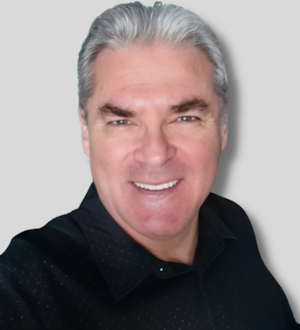 This is an image of a man wearing a black T-shirt. He is standing in front of a white background. He is looking straight ahead with a serious expression on his face. His arms are relaxed at his sides. He is wearing a form-fitting black T-shirt with a v-neck collar. His dark brown hair is combed back away from his face. His dark eyes appear intense and focused. He has a strong jawline and a thin moustache. His skin is slightly tanned. He wears a silver watch on his left arm. He looks confident and in control.