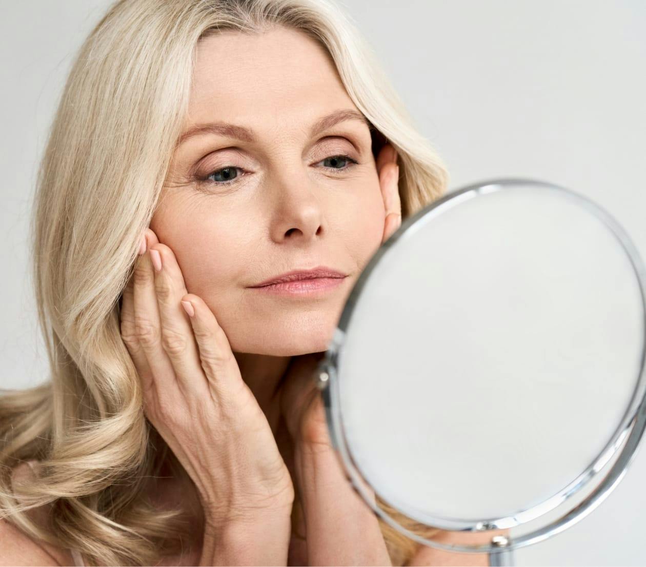 older blonde woman touching her cheeks and looking into a mirror