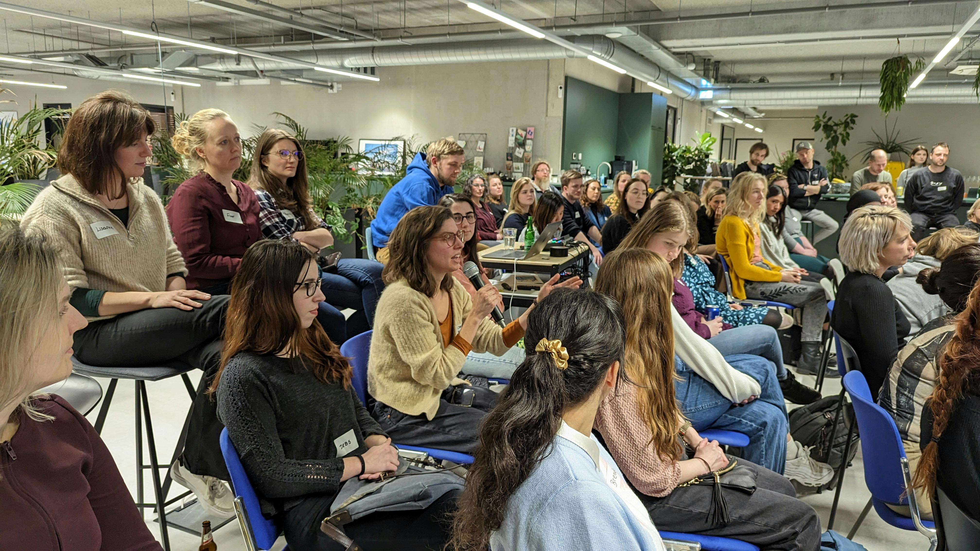 View of the audience at the Girl Code meetup hosted in 2023. An attendee sitting in the audience is holding a microphone and asking a question to the speaker.