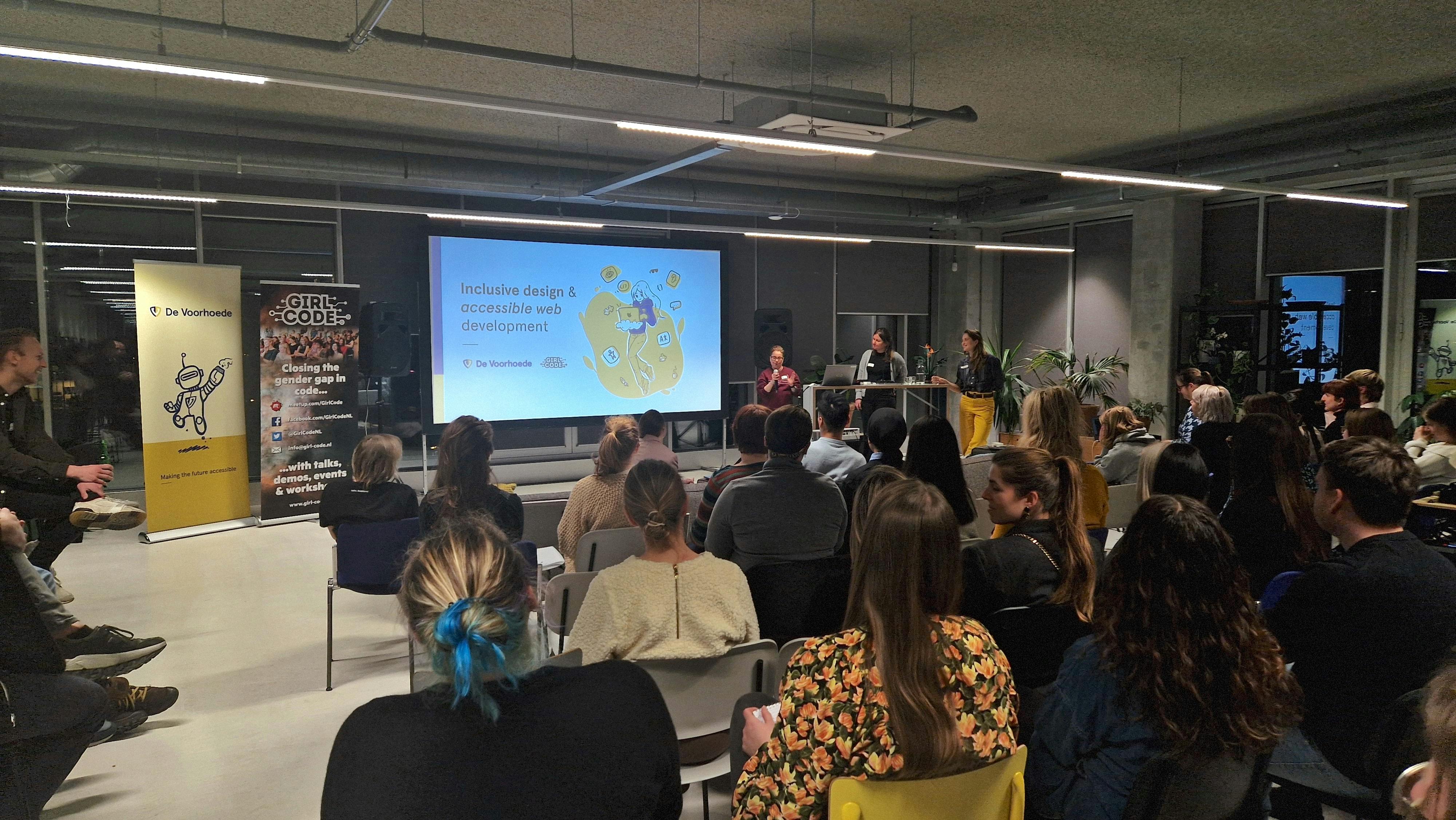 A group of people sitting in chairs listening to a woman presenting in a front of a big screen that says 'inclusive design & accessible web development.