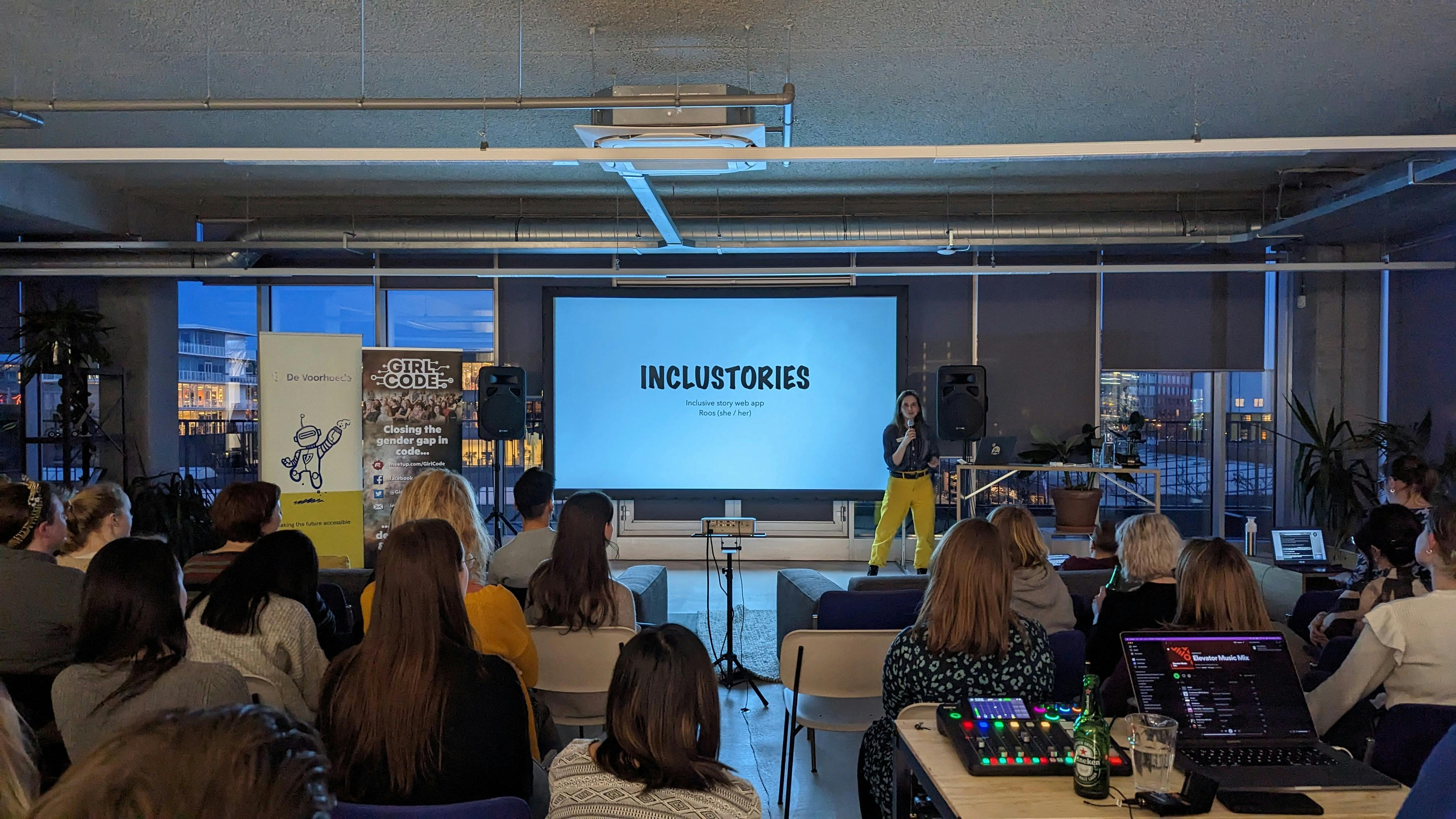 Women in a blue shirt and yellow pants presenting in front of a big screen that says 'inclustories'
