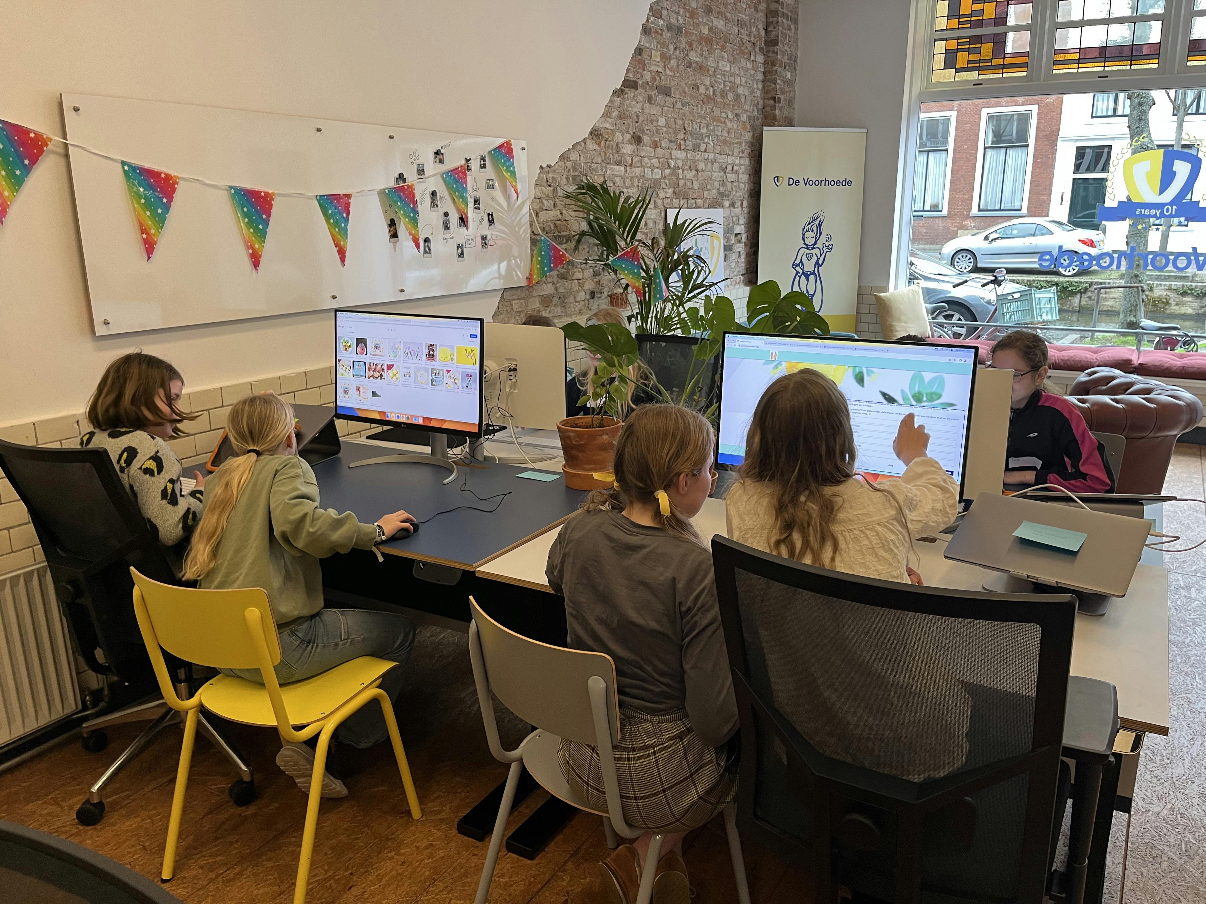 An office where four young girls are sitting behind two computers together.  They are looking at the screen and reading a question. One girl is pointing at the screen.