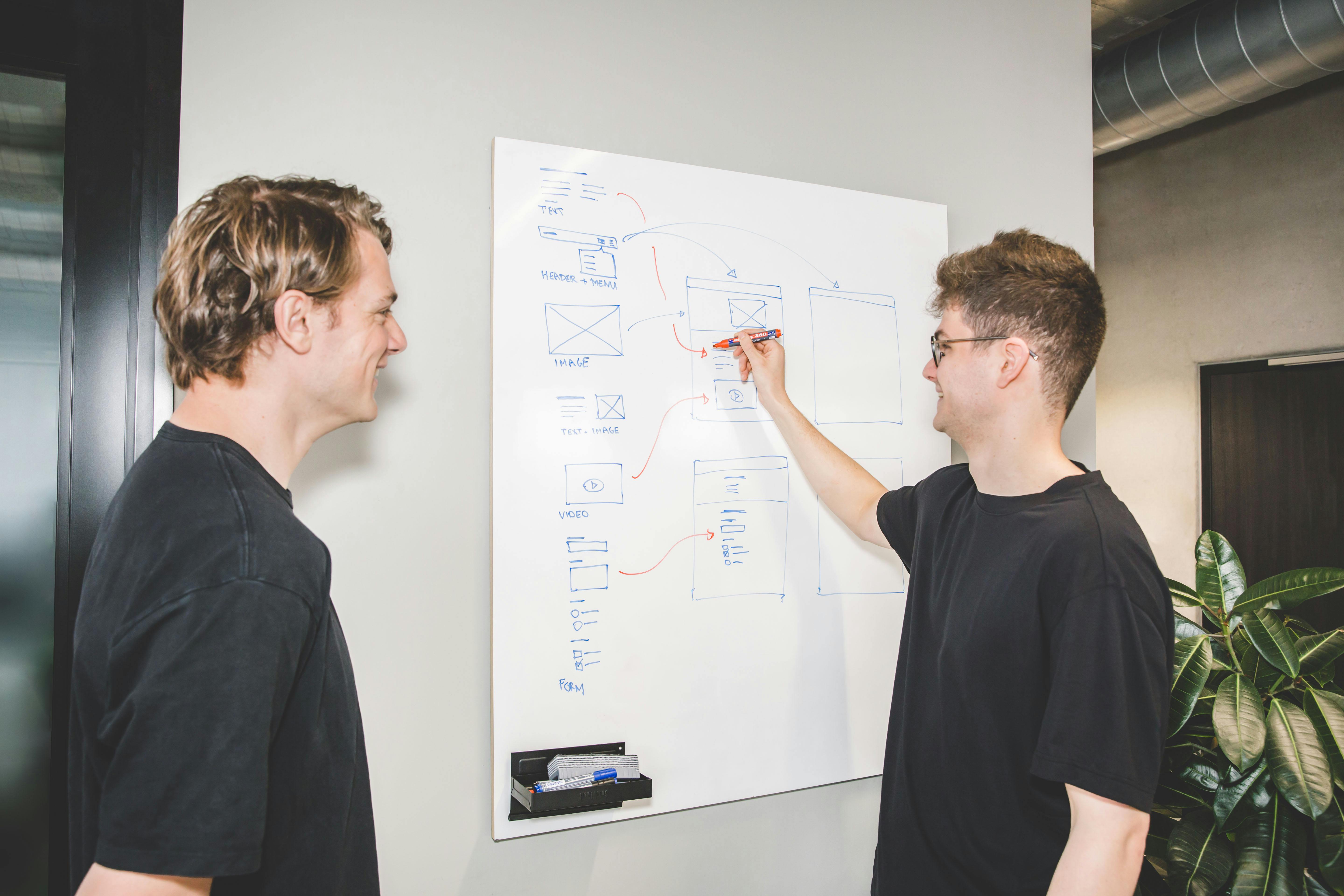 Two young men looking at a whiteboard, one is drawing a technical structure
