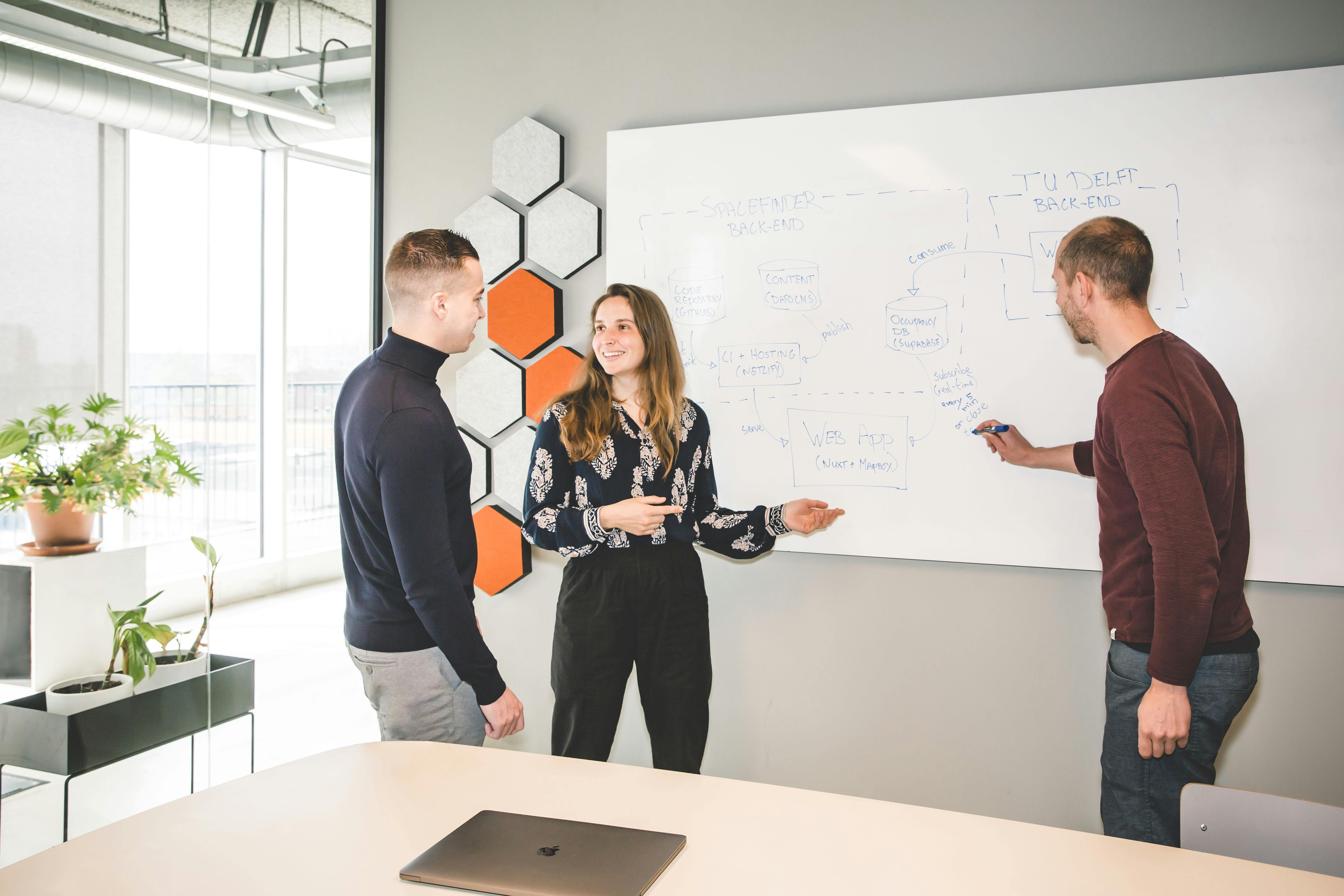 Tree people discussing a technical architecture in front of a whiteboard. A men and a woman are talking while an other men is writing on the board.