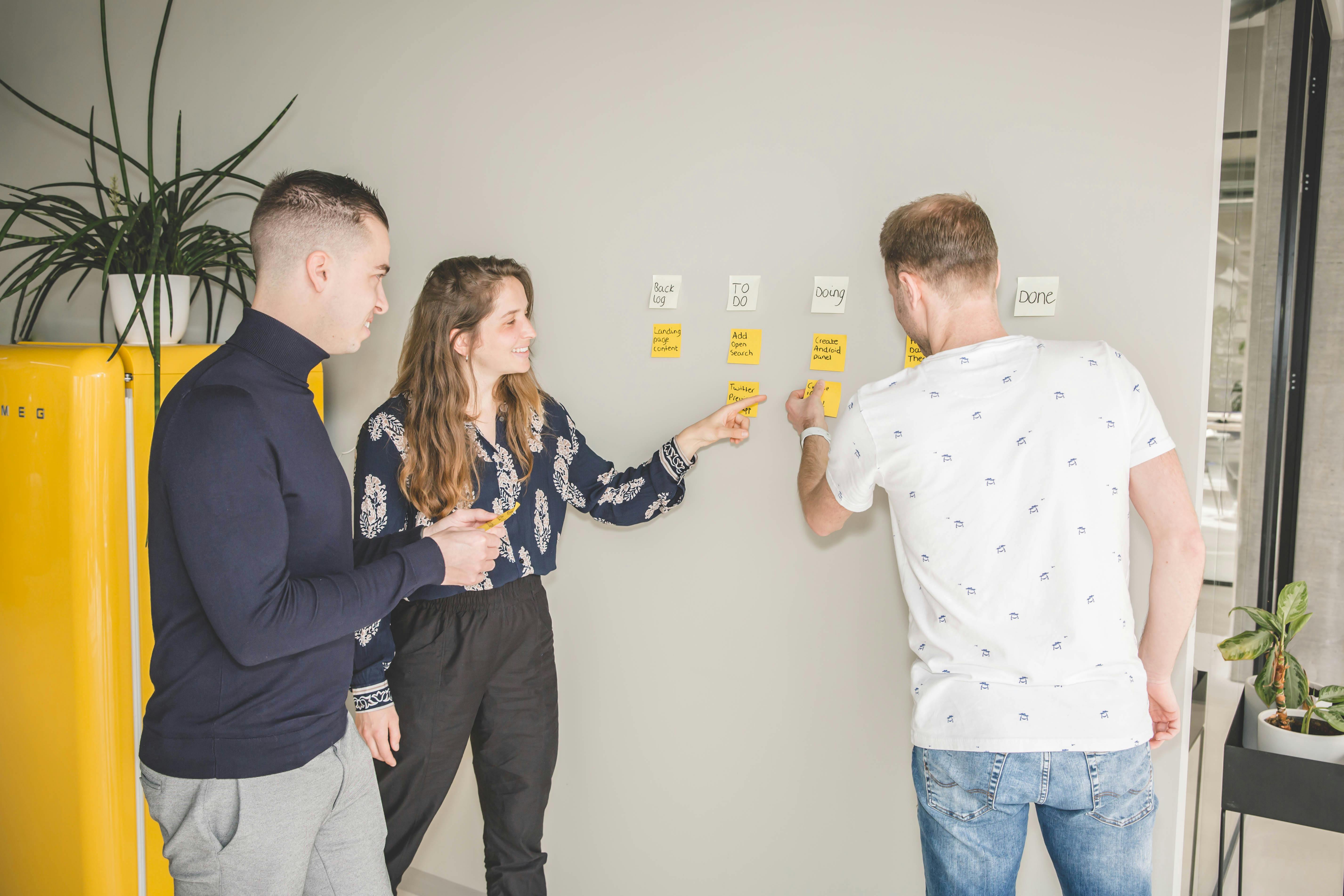 A man, a woman and another man standing at a scrumboard, holding a post-it