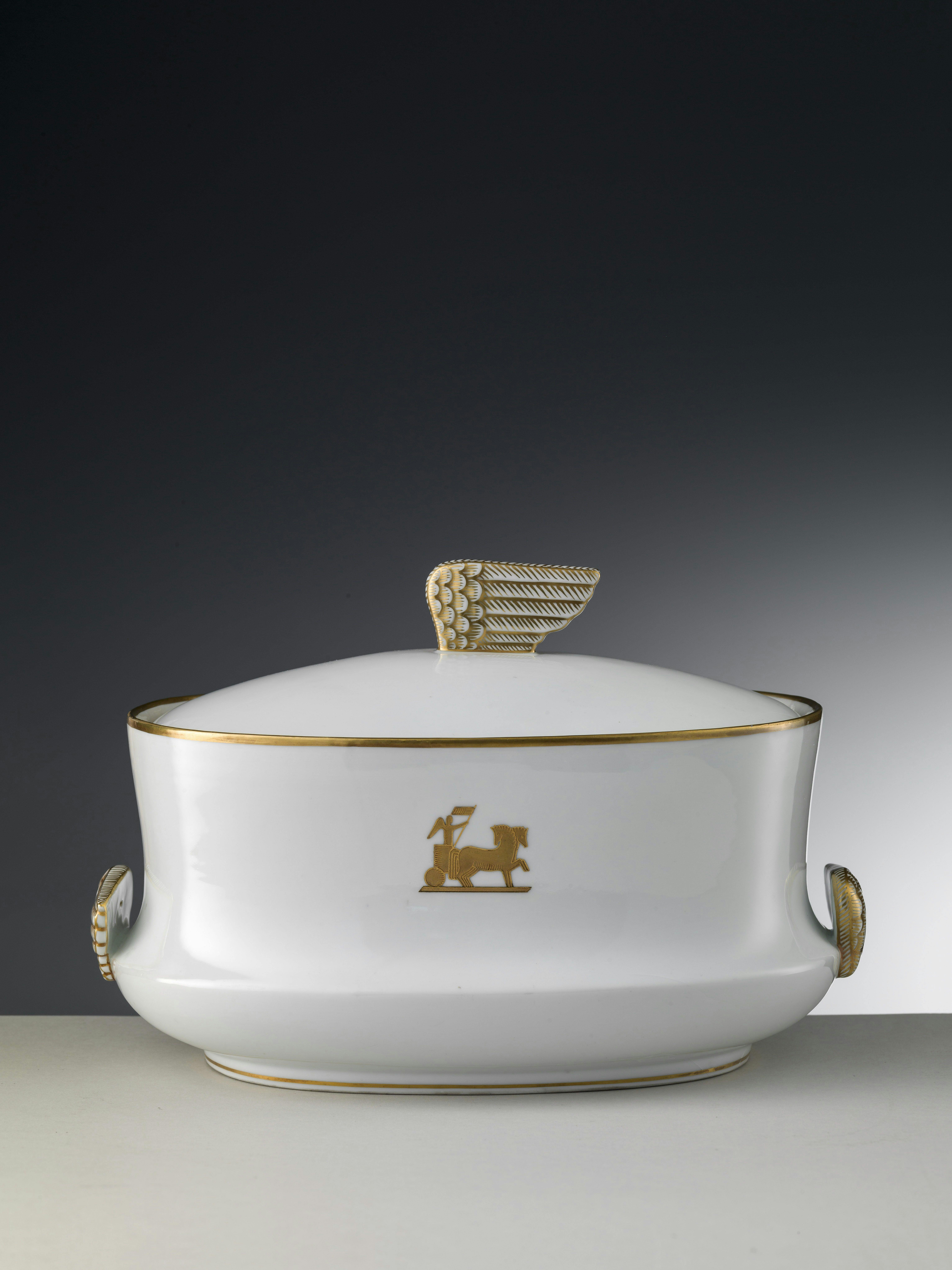 White tureen with gold rim and decorations, and a handle in the shape of a stylized wing.