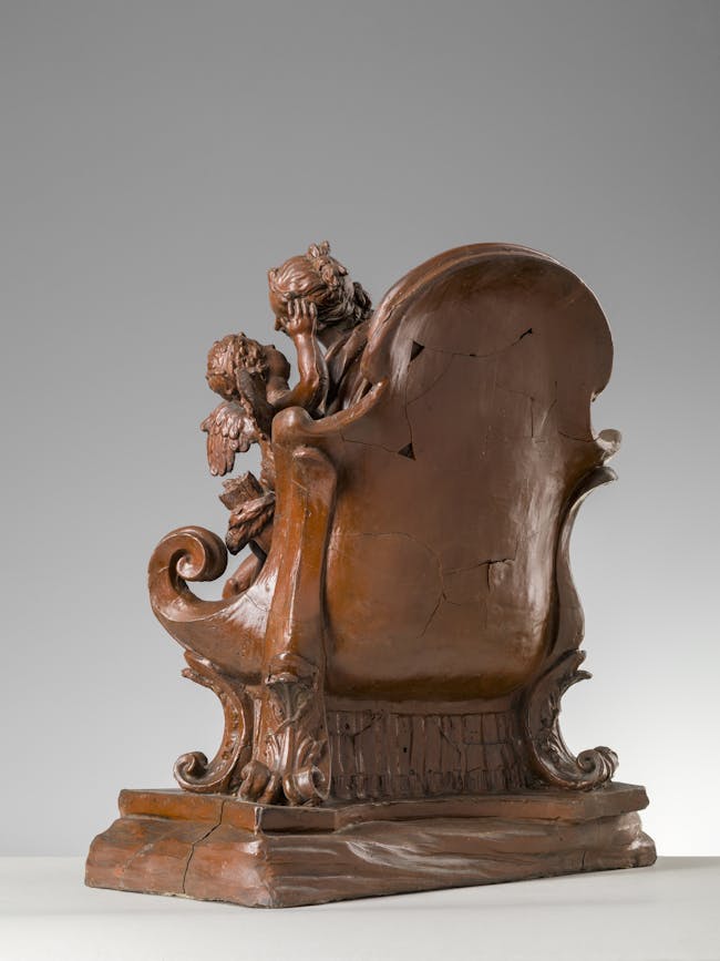 vista di tre quarti della scultura, con un seggio su cui siede una figura femminile con in braccio iun bambino