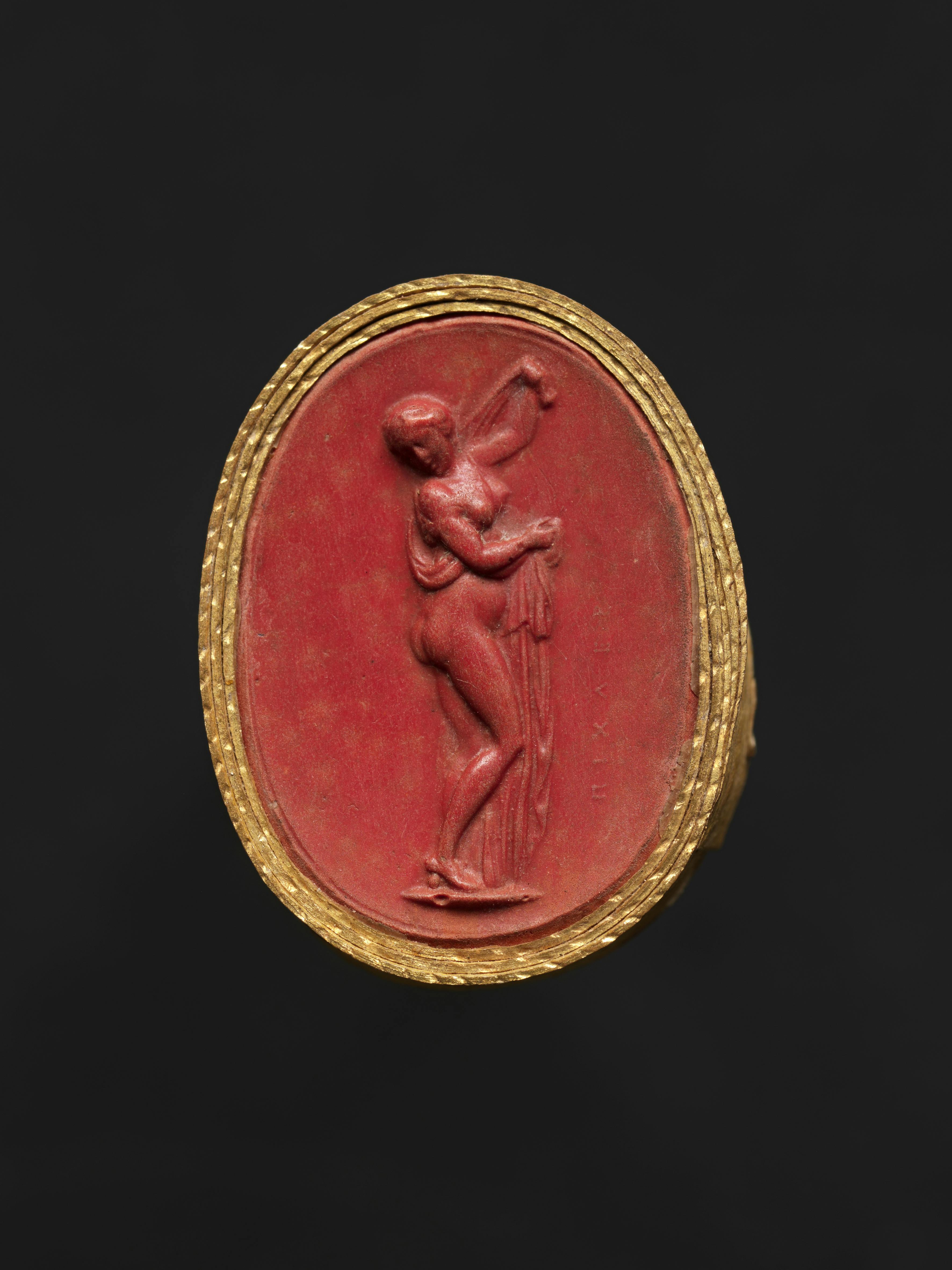 Ovale rosso con cornice dorata. Al centro è raffigurata una figura femminile con un braccio alzato.