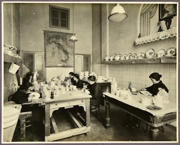 nella foto in bianco e nero sette decoratrici lavorano sedute intorno a dei tavoli di legno