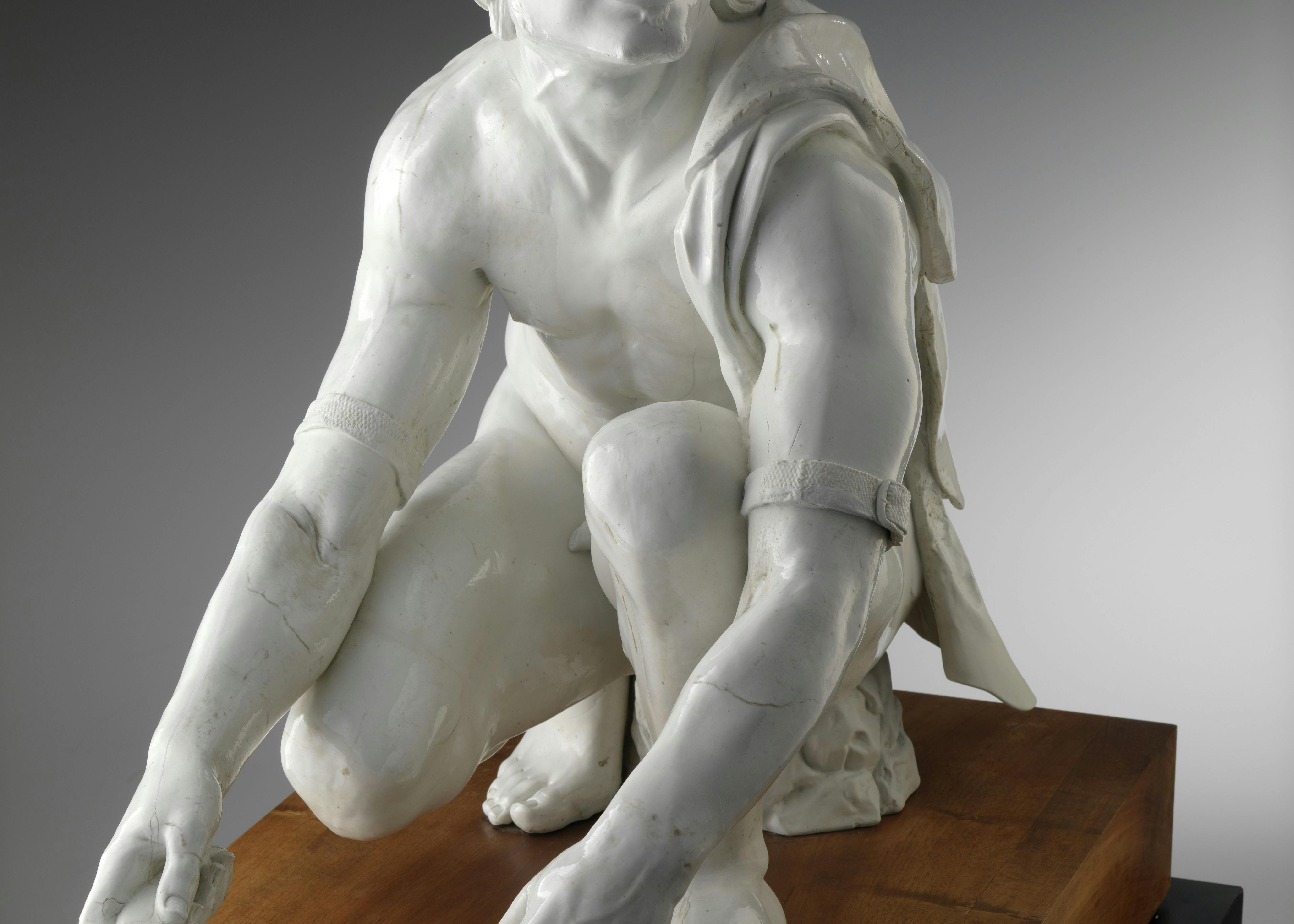 Vista frontale della scultura di porcellana bianca che raffigura un uomo inginocchiato intento a limare un coltello