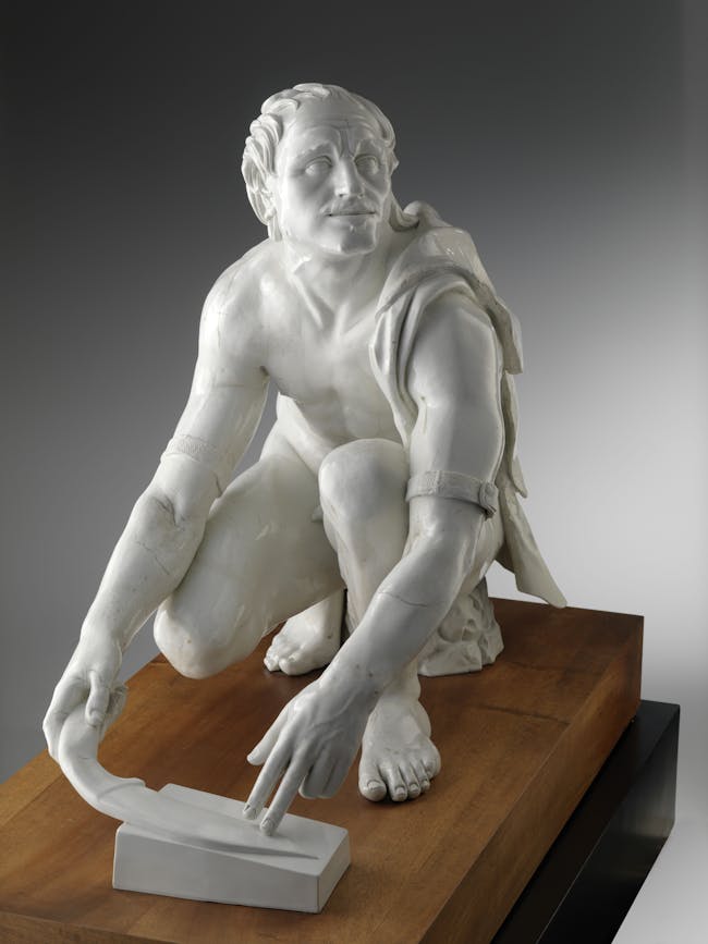 Vista frontale della scultura di porcellana bianca che raffigura un uomo inginocchiato intento a limare un coltello