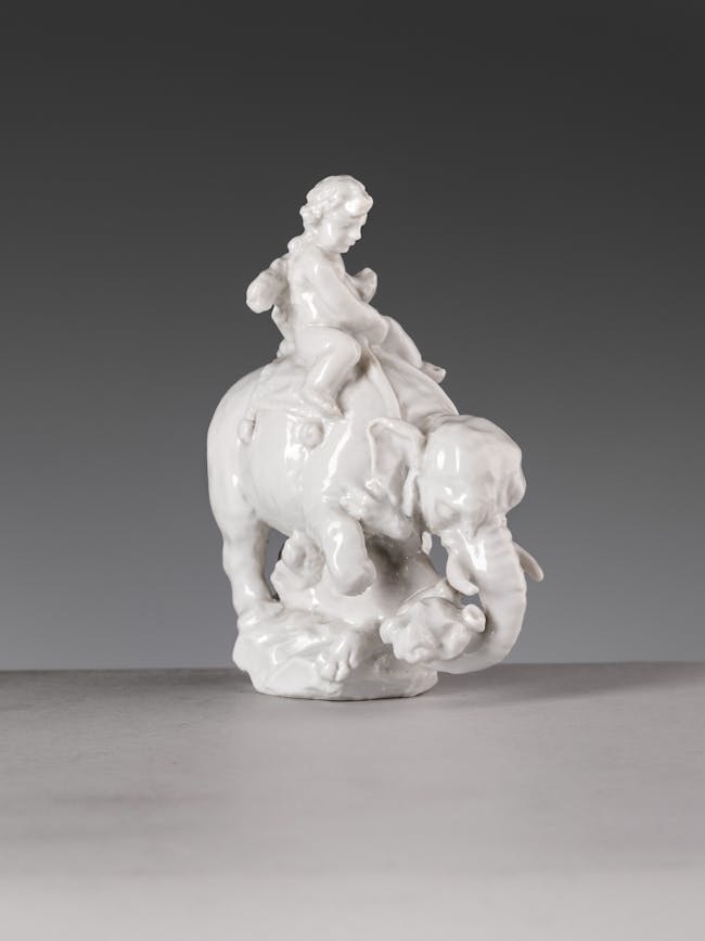 Piccola scultura di porcellana bianca raffigurante un bambino a cavallo di un elefante
