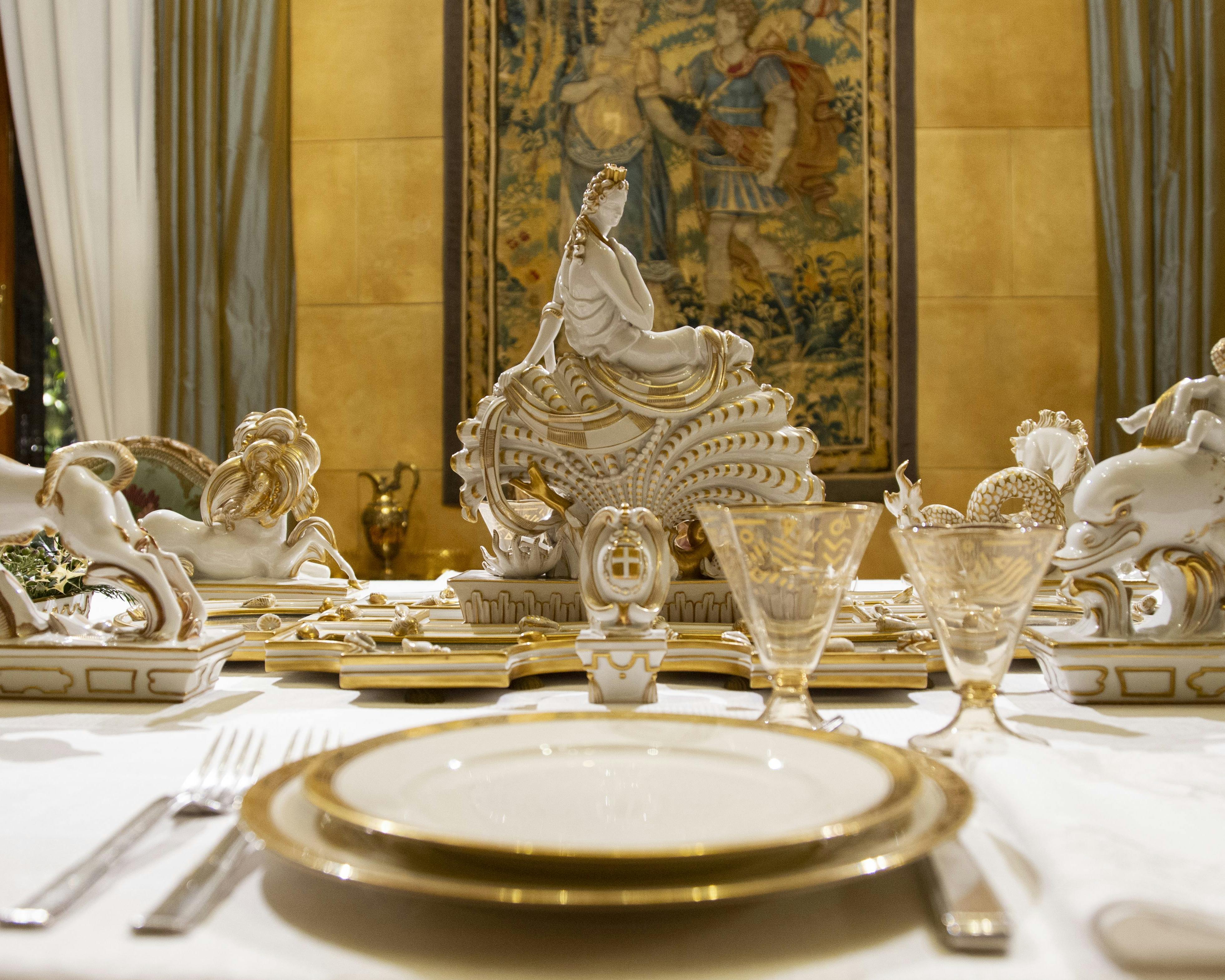 In primo piano un piatto bianco e oro, sullo sfondo statue in porcellana per il decoro della tavola