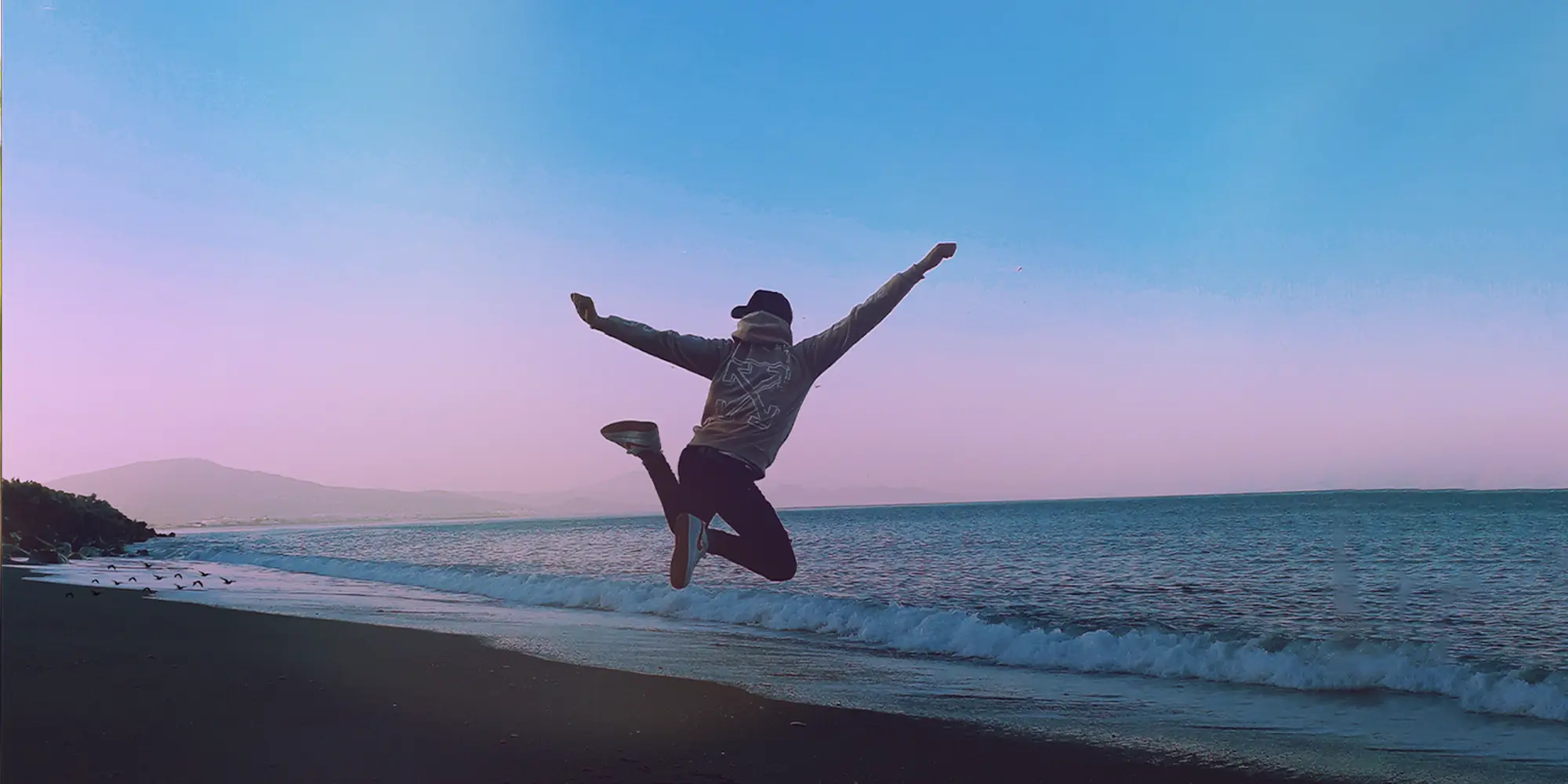 Imagem com homem saltando com braços abertos e pernas flexionadas em frente ao mar.