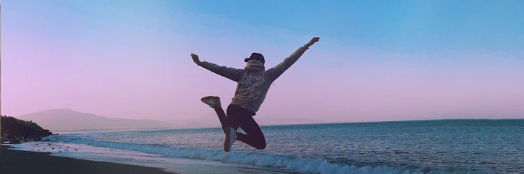 Imagem com homem saltando com braços abertos e pernas flexionadas em frente ao mar.