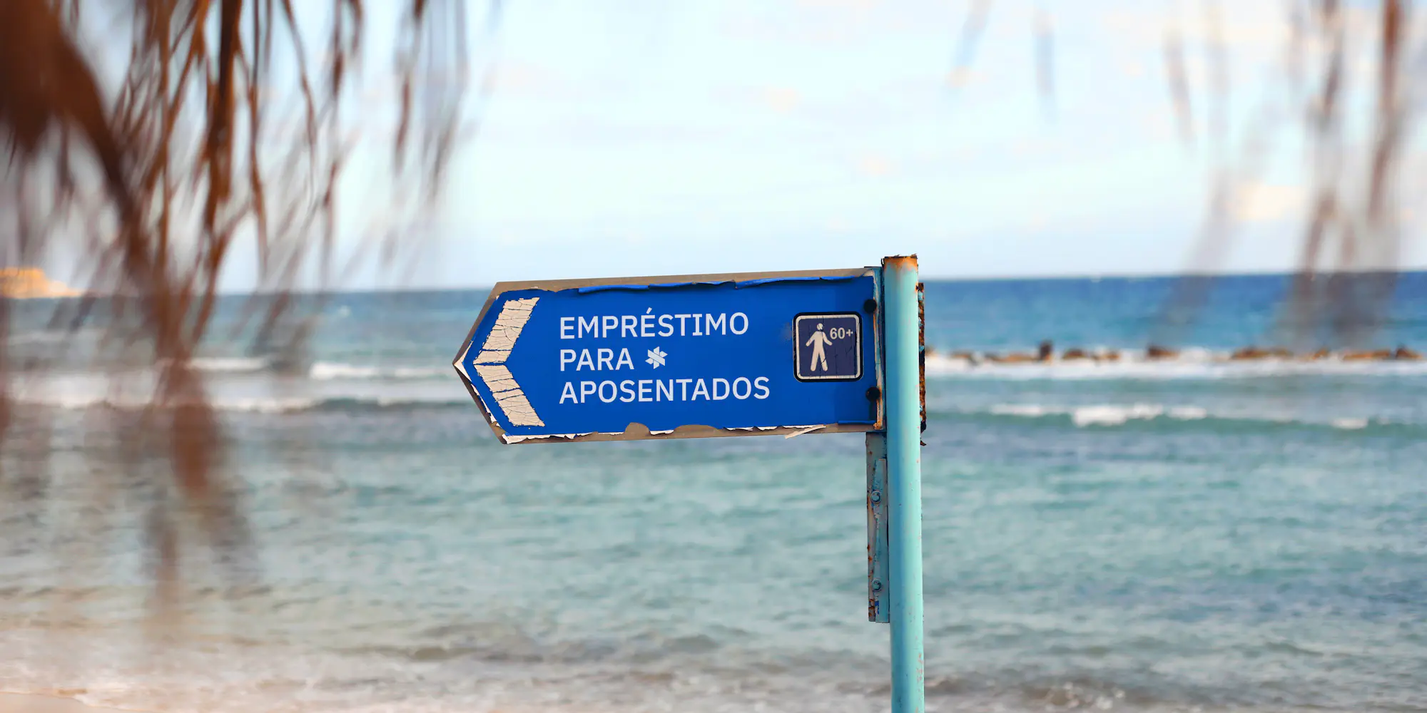 Placa de direção azul fincada na areia da praia com mar ao fundo.