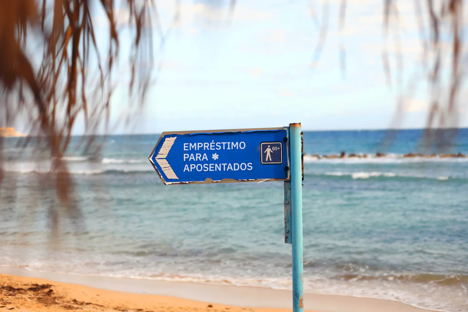 Placa de direção azul fincada na areia da praia com mar ao fundo.