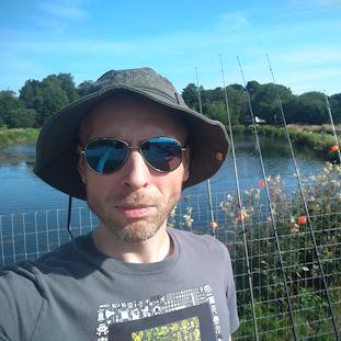 A self-portrait of Game Director, Paul "Rushy" Rustchynsky. Background: a fence, lined with fishing rods, in front of a small lake.