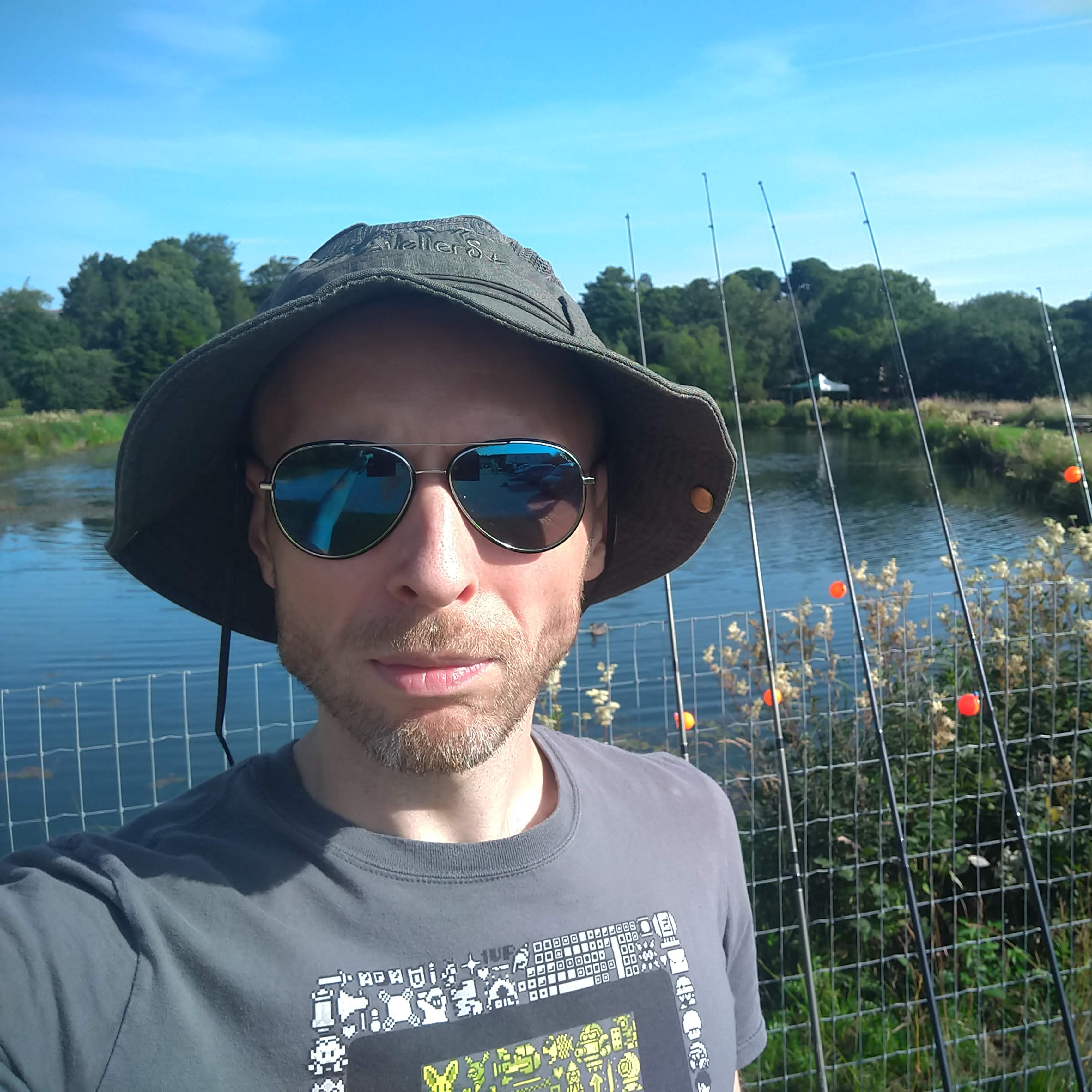 A self-portrait of Game Director, Paul "Rushy" Rustchynsky. Background: a fence, lined with fishing rods, in front of a small lake.