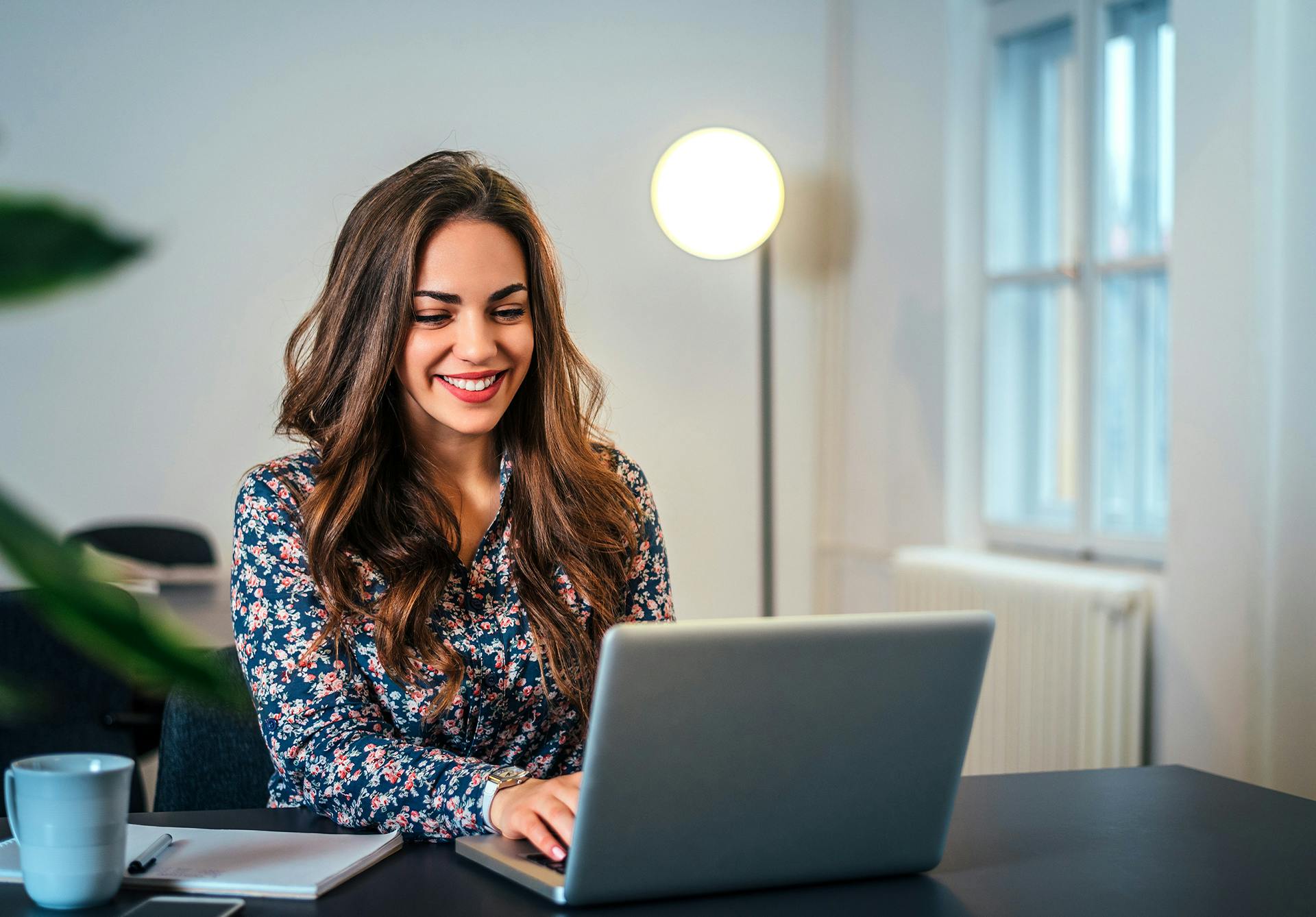 brunette woman sitting at a desk on her laptop, with a coffee mug next to her