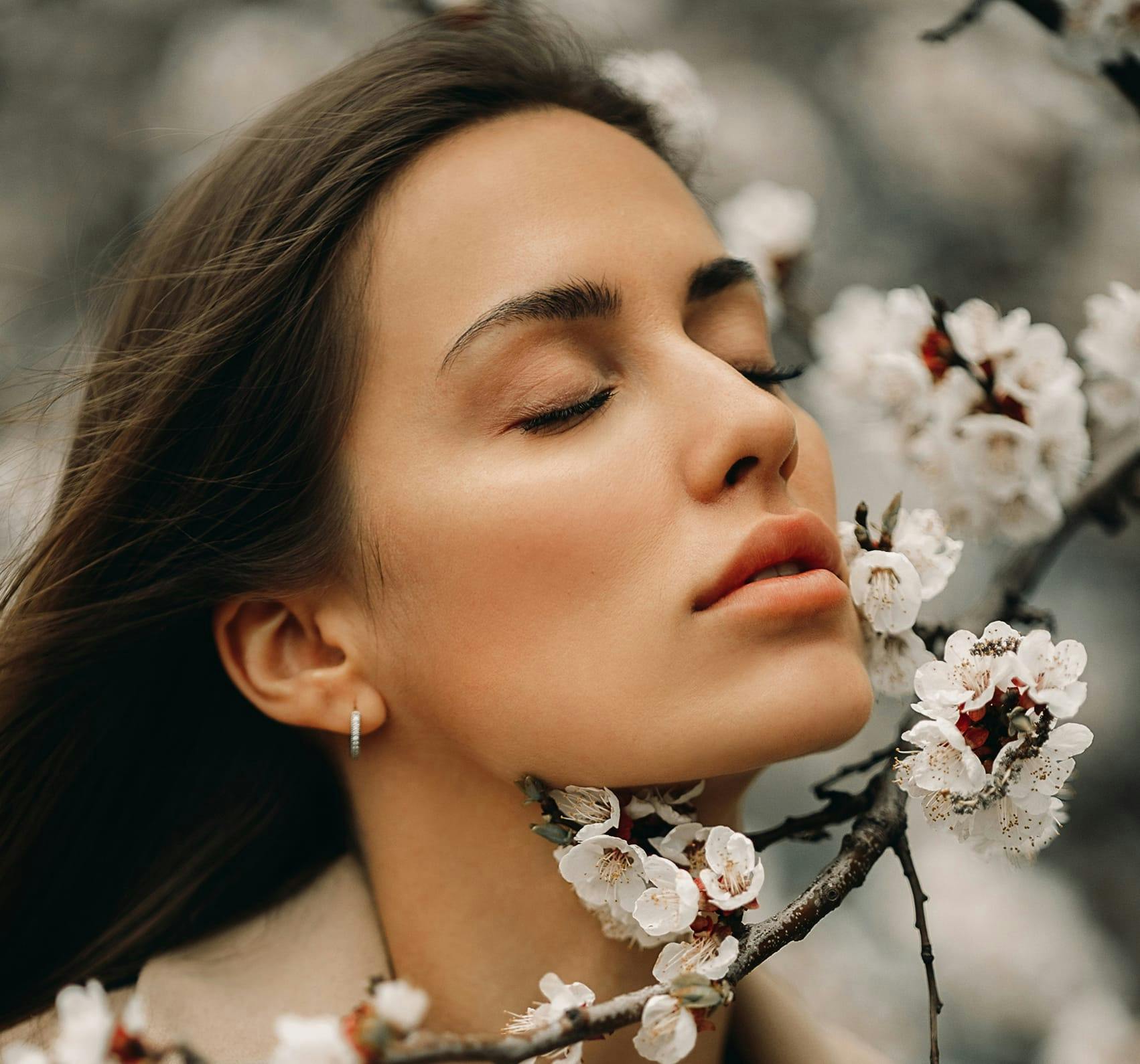 A dark haired woman with clear skin smelling flowers.