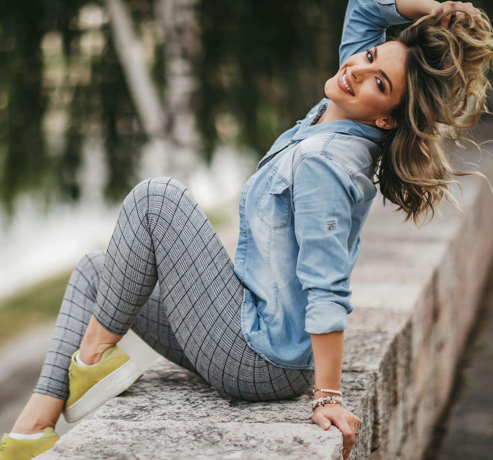A blonde woman sitting on a stone wearing plaid pants & a blue jean jacket.