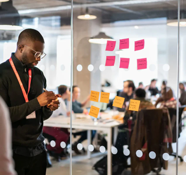 Image of a man writing post it note, people in an office in the background