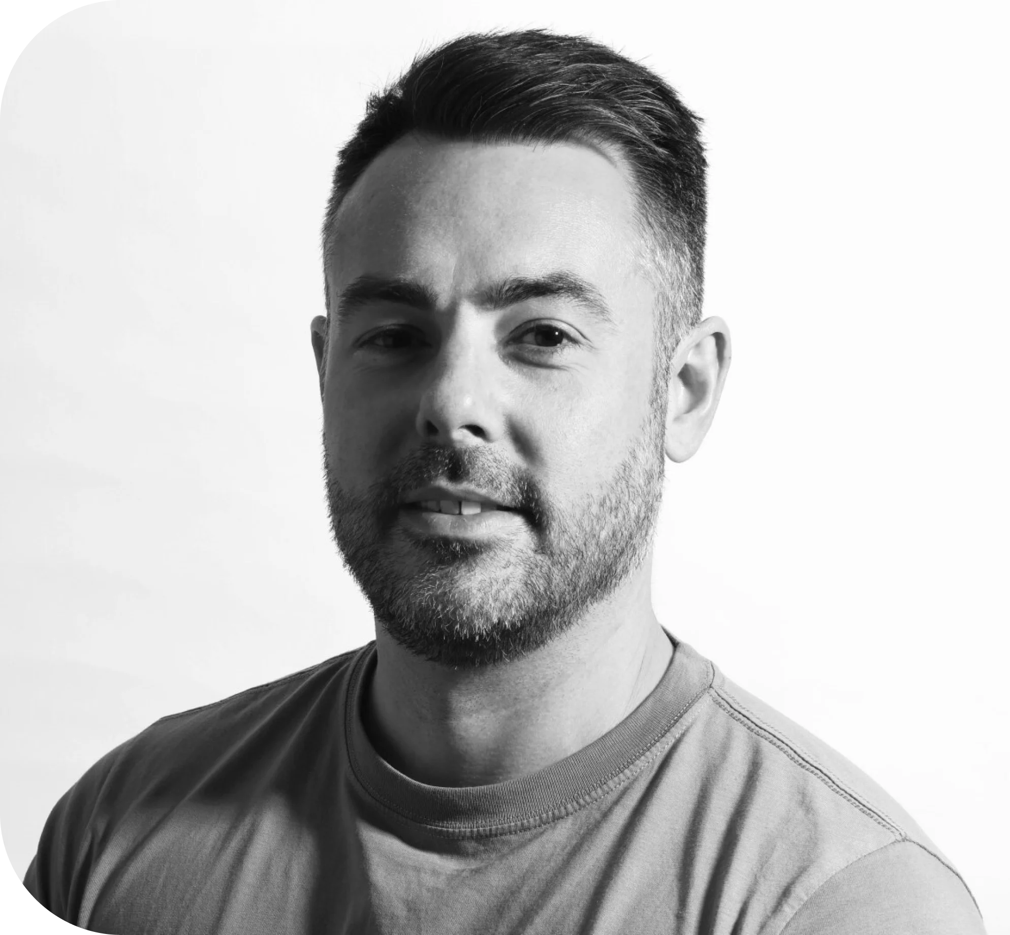 Black and white portrait of a man with short dark hair and a trimmed beard, wearing a crew-neck t-shirt, smiling slightly and looking towards the camera, set against a plain light background.
