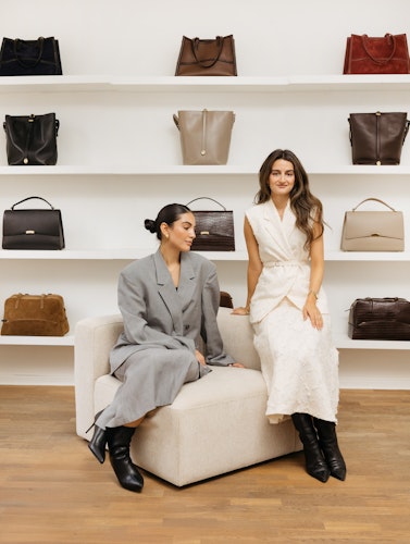 de to grundlæggere i en lænestol i deres lyse og luftige showroom med et udvalg af deres håndtasker på væggen bag dem