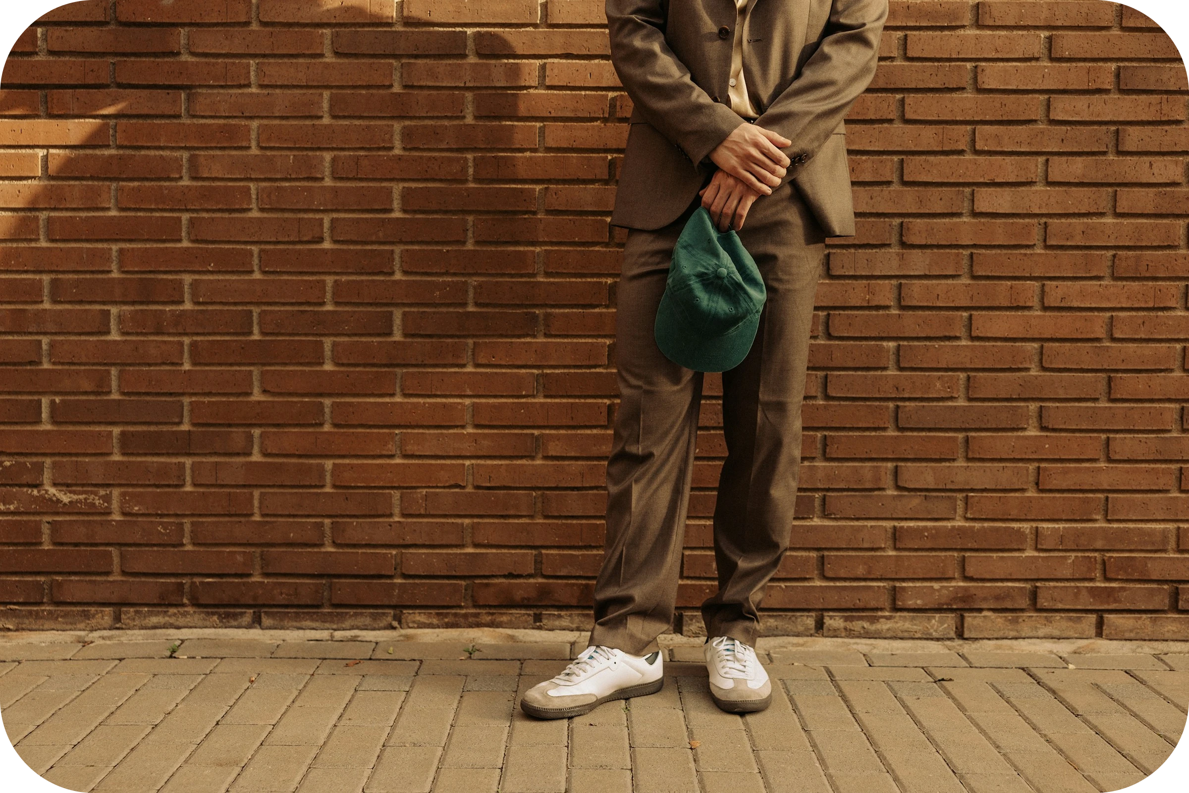 Rennosti pukeutunut mies seisoo tiiliseinää vasten ja pitää kädessään vihreää lippalakkia.