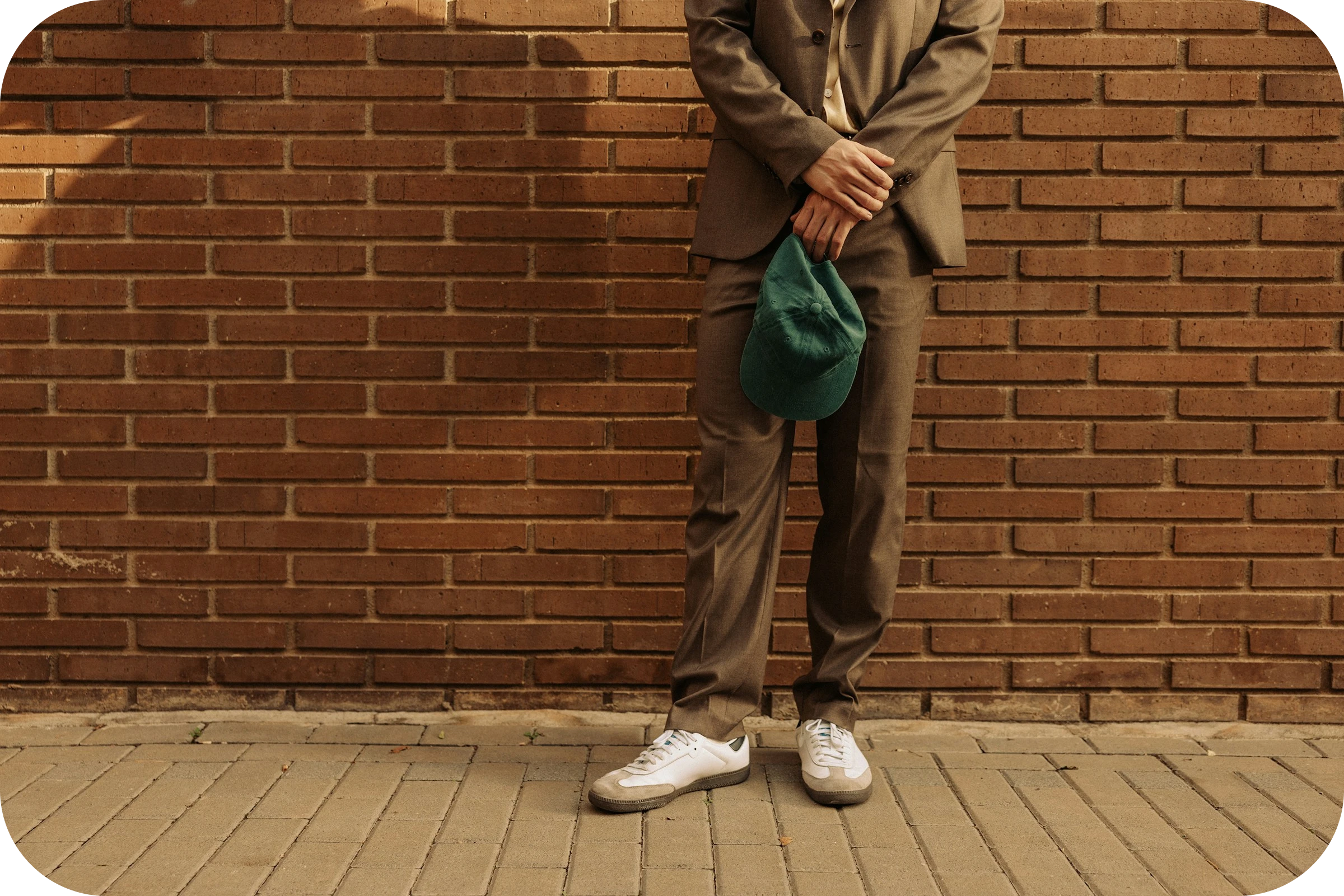 Rennosti pukeutunut mies seisoo tiiliseinää vasten ja pitää kädessään vihreää lippalakkia.