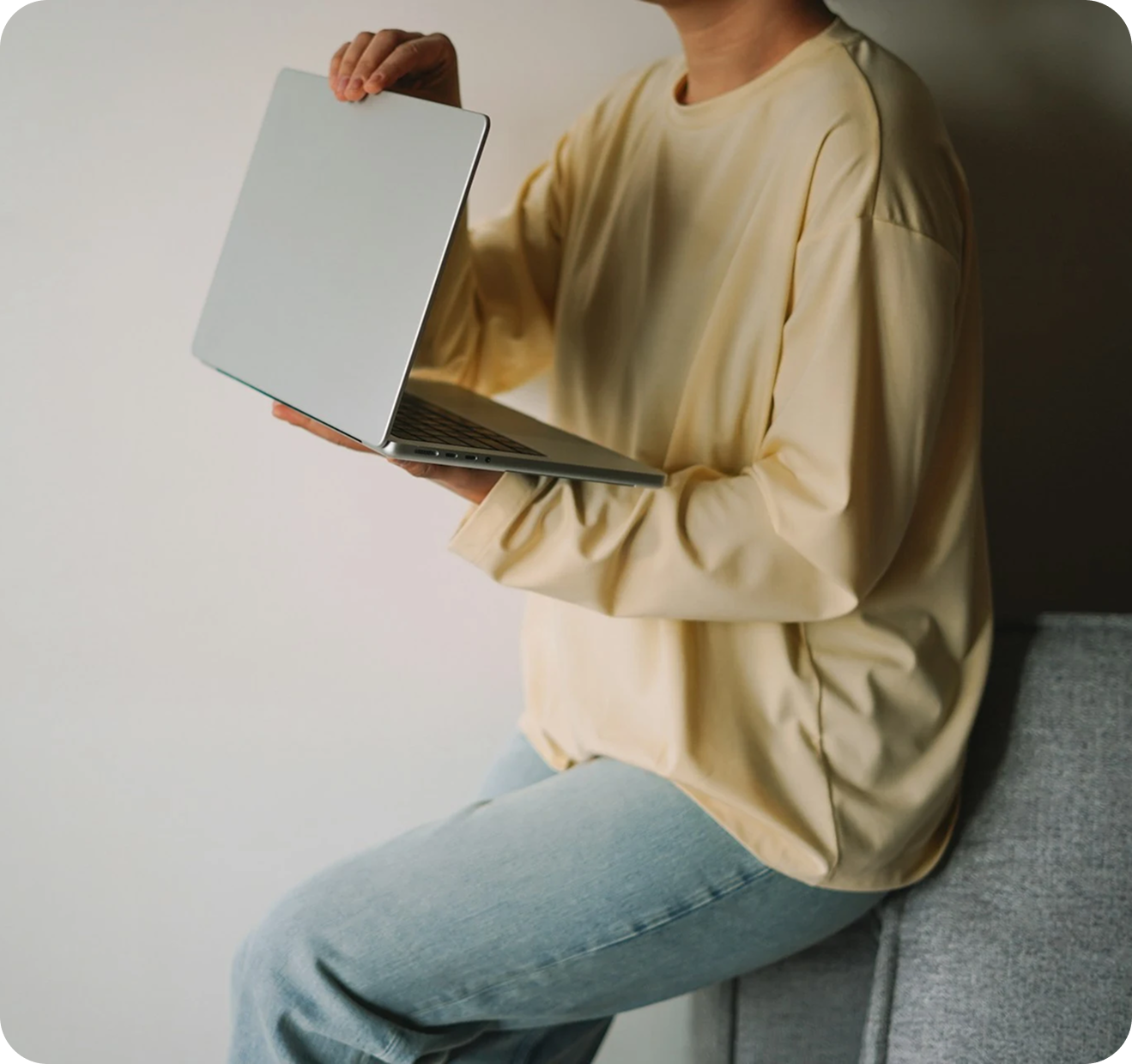 person sitting at the couch end holding a laptop and wearing a relaxed outfit in light colors