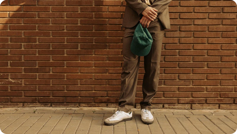 Image with cool unexpected vibe that reads more as fintech than a bank, the image shows a person leaning casually against brick wall holding green cap in hand and wearing a casual brown suit and white sneakers