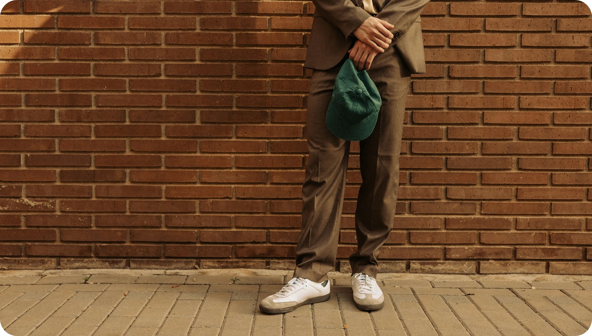 Image with cool unexpected vibe that reads more as fintech than a bank, the image shows a person leaning casually against brick wall holding green cap in hand and wearing a casual brown suit and white sneakers