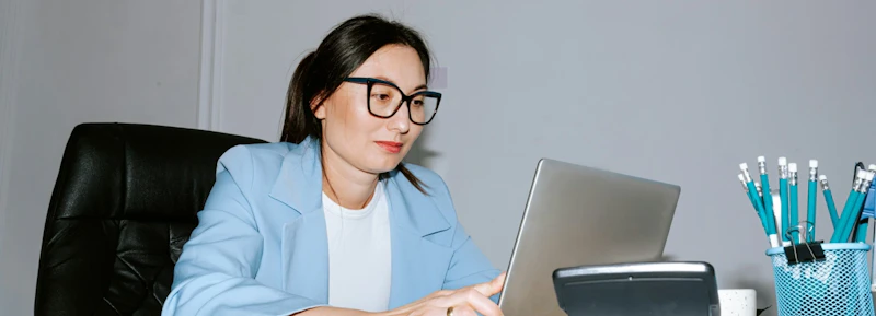 Woman with t-shirt, light blue sports blazer and glasses focusing on something on her laptop screen