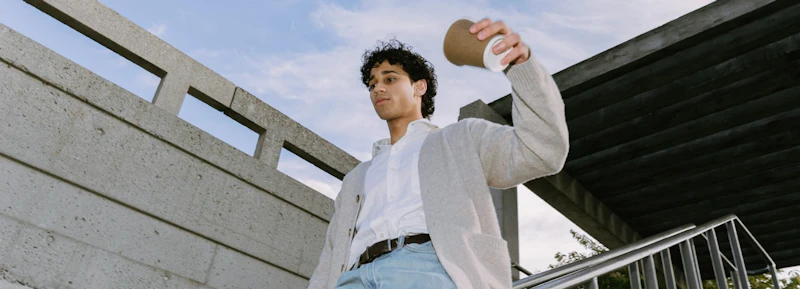 Man with dark curly hair holding a coffee cup close to his face while walking down the stairs outside