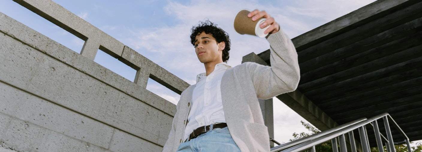 Man with dark curly hair holding a coffee cup close to his face while walking down the stairs outside