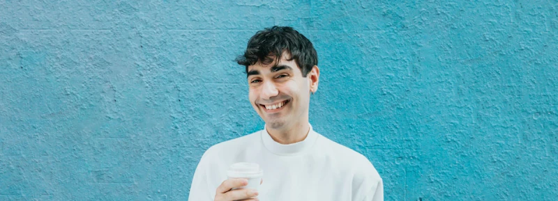Man with dark hair standing against a blue wall smiling at the camera while holding a coffee