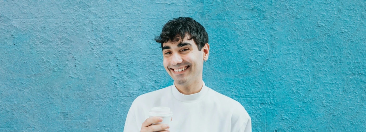 Man with dark hair standing against a blue wall smiling at the camera while holding a coffee