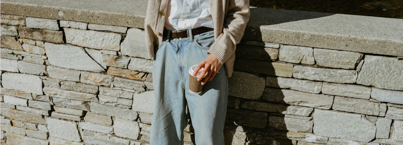 Person standing by a stone wall wearing jeans holding a coffee cup