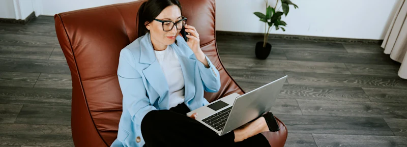Woman with professional and casual look sitting in leather chair looking at laptop screen while talking on the phone