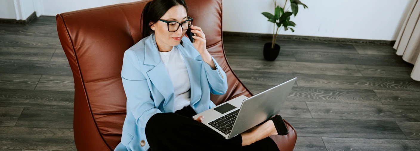 Woman with professional and casual look sitting in leather chair looking at laptop screen while talking on the phone