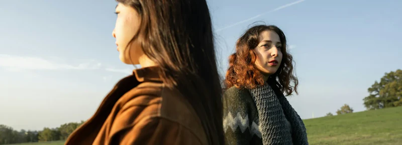two women looking in opposite directions, standing on grass field, considering their options
