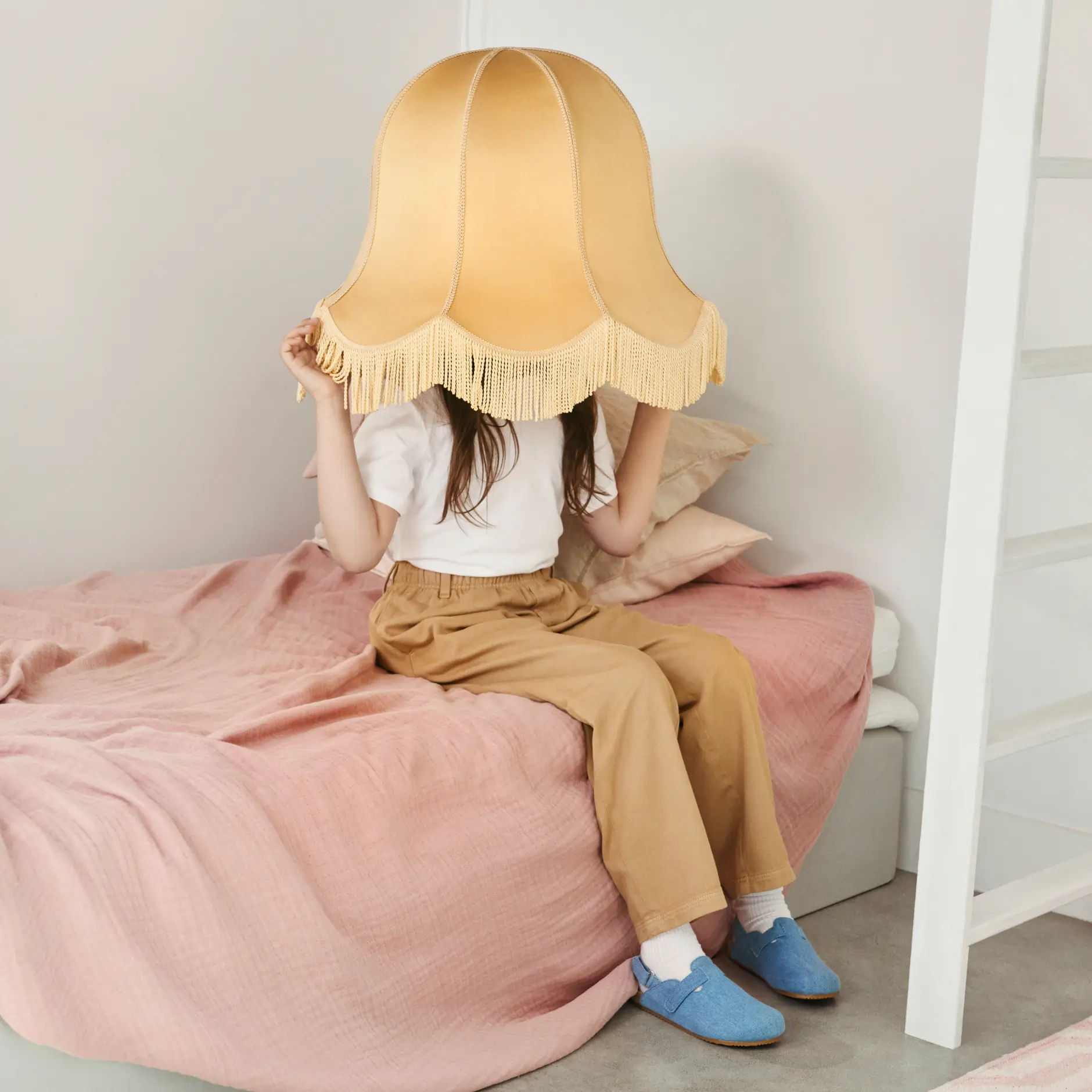 A child sits on a bed with a large yellow lampshade covering their head, styled with warm colors from Adressändring’s palette.