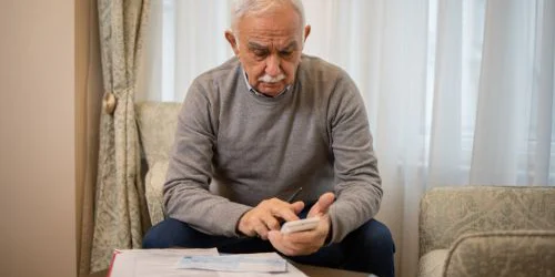 Older man sitting on couch in living room with papers in front of him and calculator in-hand